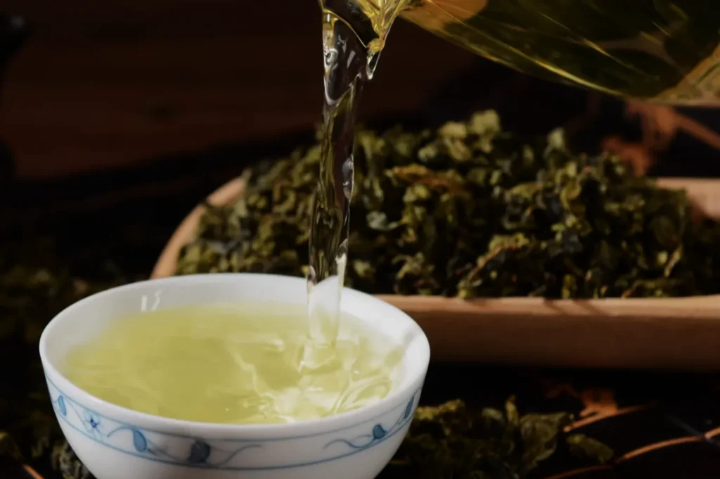 A stream of hot green tea pours into a blue-and-white porcelain cup, with loose tea leaves behind it, creating a warm, calming scene.