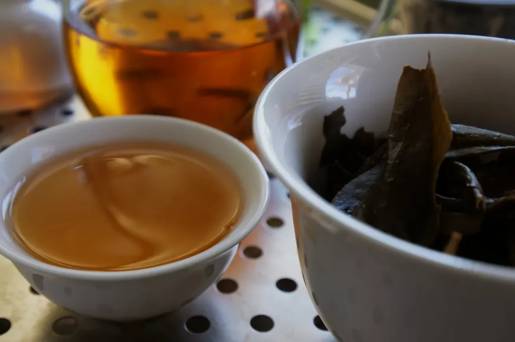 A shallow bowl of tea next to a cup containing tea leaves, with a larger pot of tea in the background.