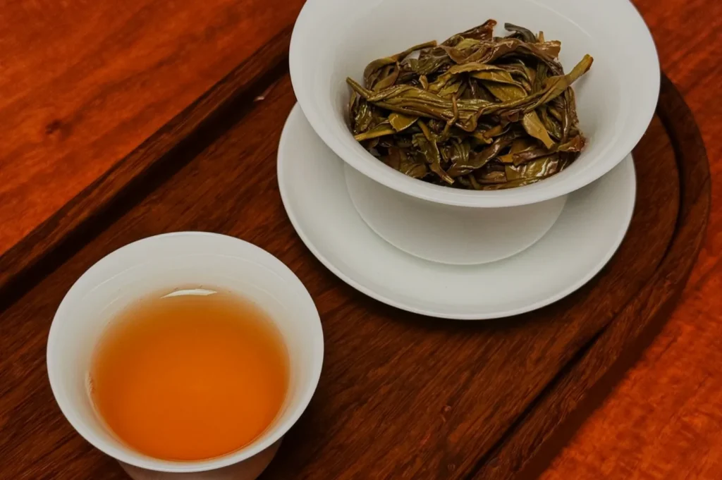 A small cup of tea next to a white bowl filled with tea leaves, all placed on a wooden tray.