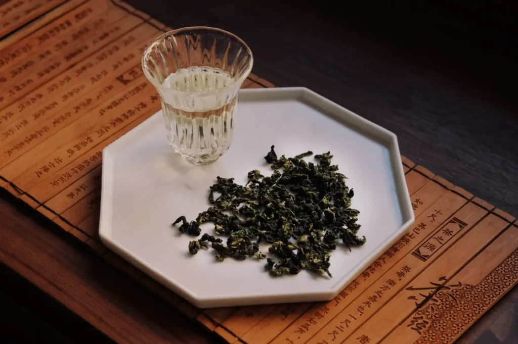 A glass of tea beside a plate of loose green tea leaves on a wooden surface with text in the background.