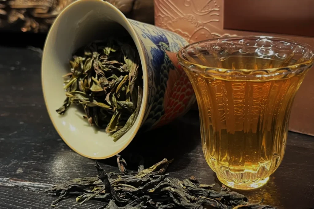A close-up of loose tea leaves spilling from a decorative cup beside a filled glass of amber tea on a wooden surface.