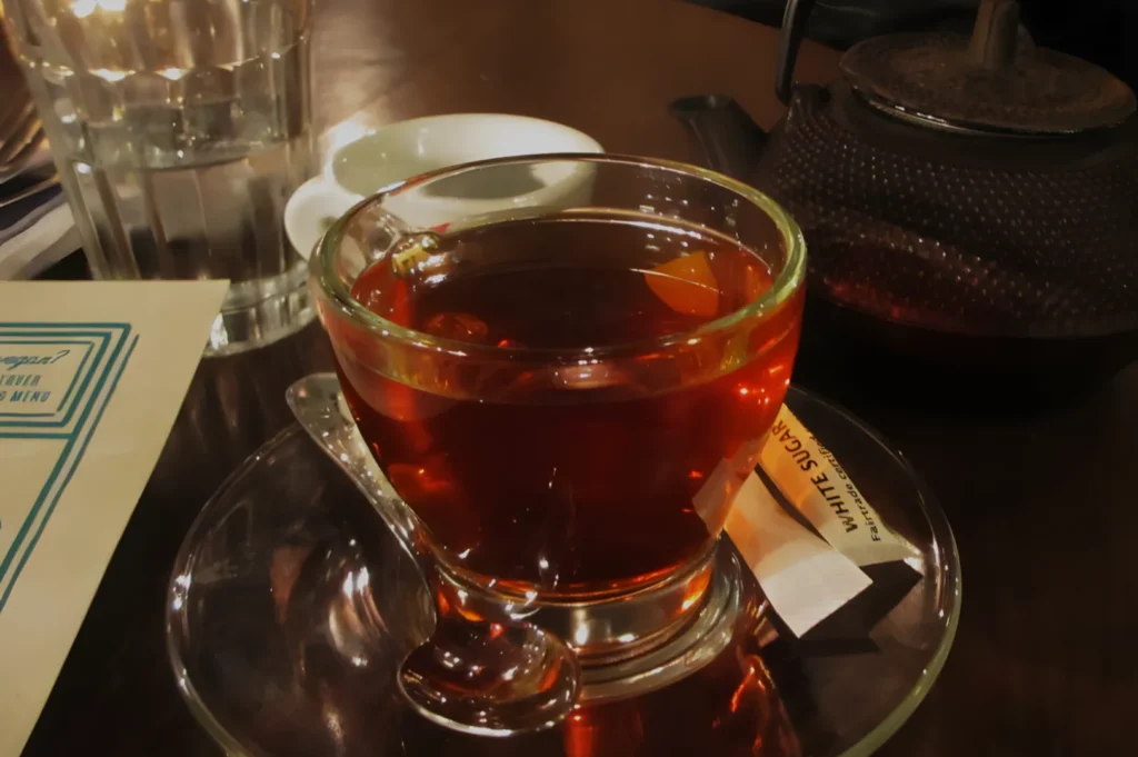 A cup of tea on a saucer with a spoon, surrounded by a glass of water and a teapot in a dimly lit setting.