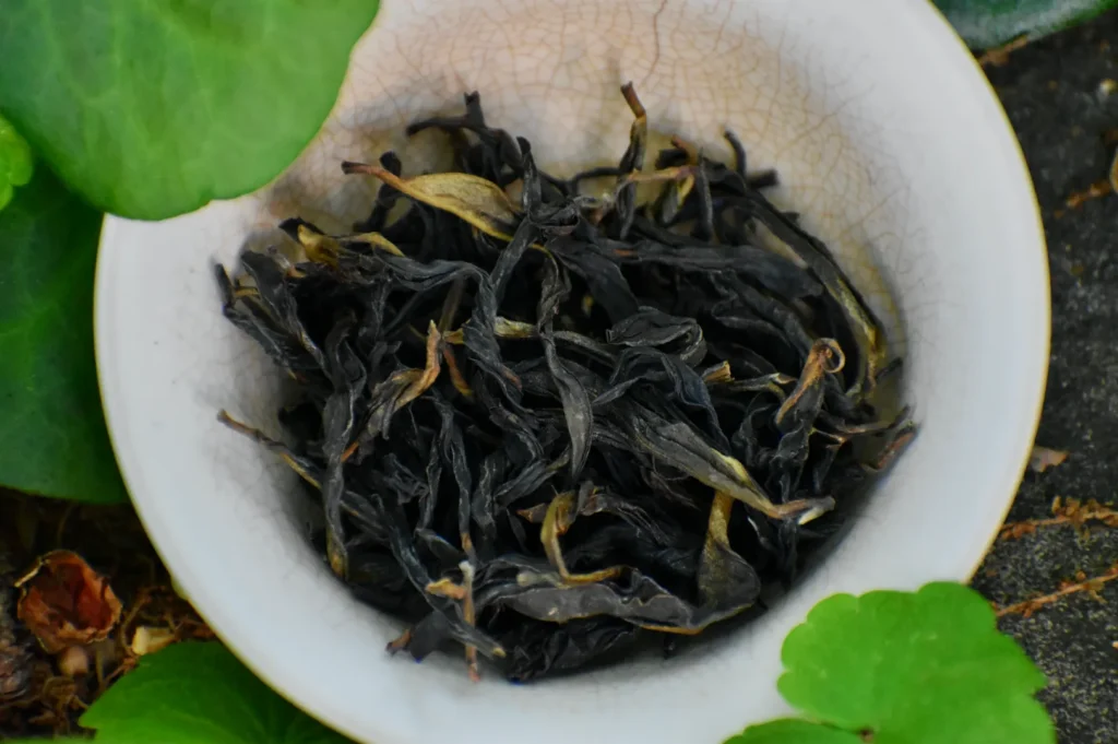 Dried black tea leaves in a small white bowl surrounded by greenery.