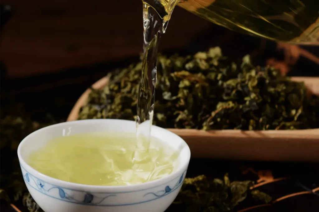 Golden-green tea is poured from a glass vessel into a white porcelain cup with blue designs, surrounded by loose tea leaves, creating a warm, serene mood.