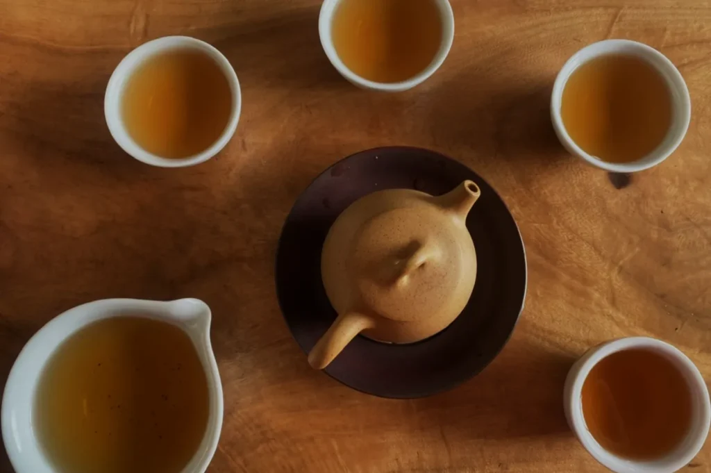 Overhead view of a round clay teapot and five cups of amber tea on a warm wooden table, creating a cozy, inviting atmosphere.