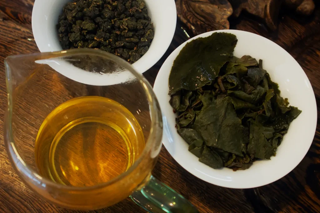 A glass teapot containing yellow tea, a bowl of dried tea leaves, and a bowl of wet tea leaves on a wooden surface.