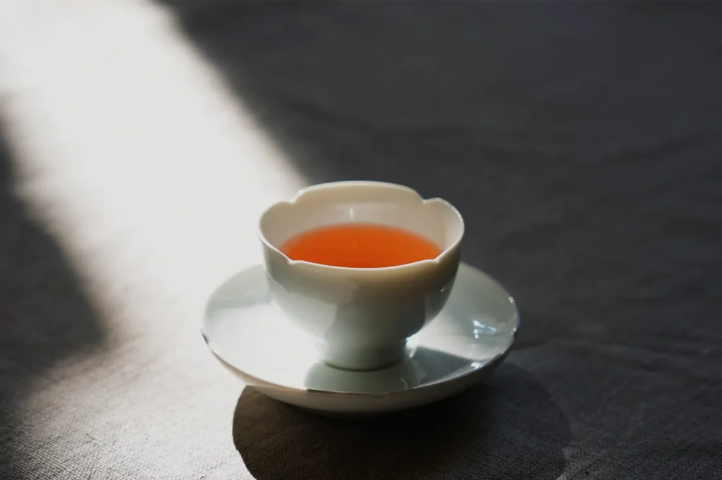 White porcelain teacup filled with amber tea rests on a matching saucer, illuminated by a sunbeam on a dark table, creating a calm mood.
