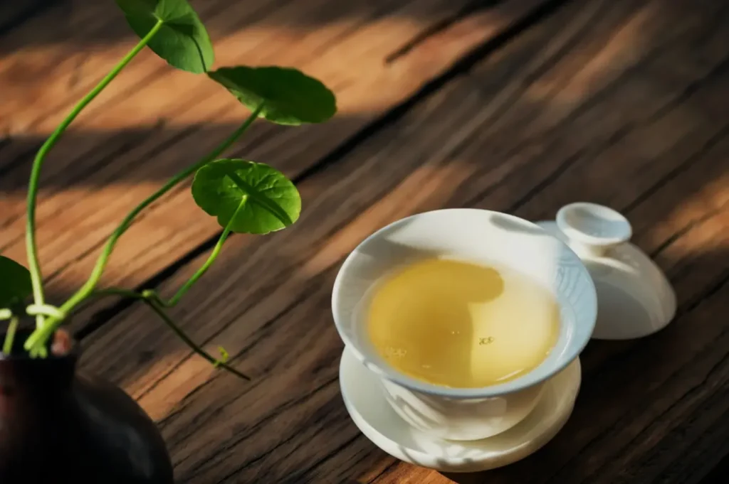 A cup of light-colored tea on a wooden surface, accompanied by a small teapot and a green plant in the background.
