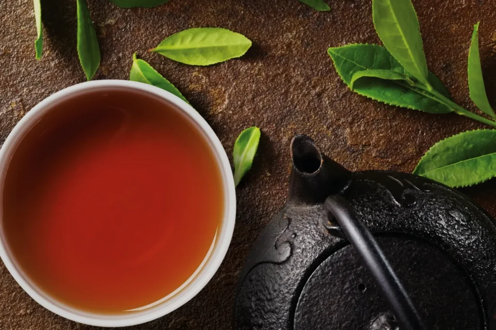 A ceramic cup of tea next to a cast iron teapot and fresh tea leaves on a textured surface.