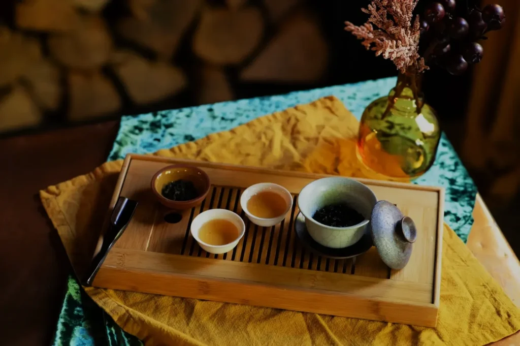 A tea set arranged on a wooden tray, featuring multiple small cups with tea, a teapot, and a decorative vase with dried flowers and grapes in the background.