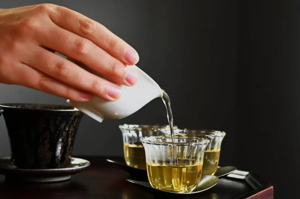 A hand pouring tea from a small white vessel into three clear tea glasses on a dark background.