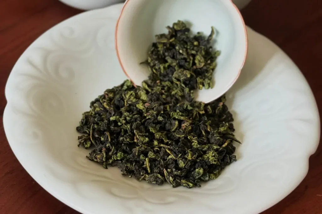 Dried green tea leaves on a white plate with a small bowl tipped over, spilling more leaves.
