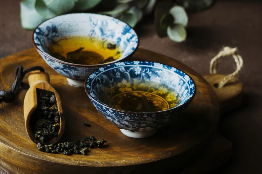 Two blue and white patterned bowls of tea on a wooden board, accompanied by a wooden scoop filled with loose tea leaves.