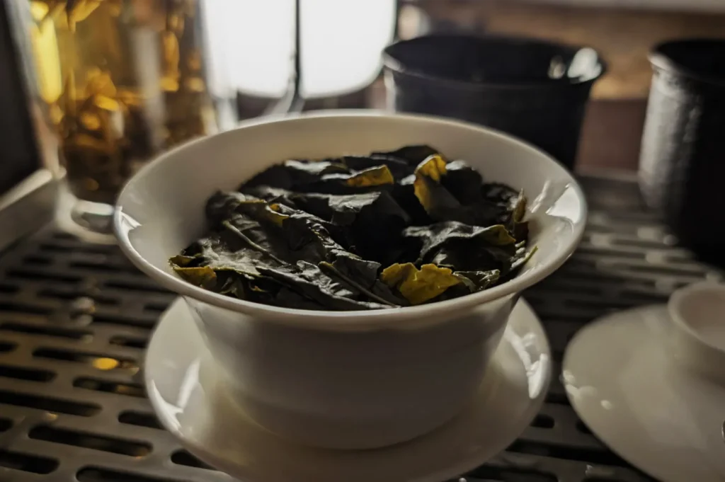A white ceramic bowl holds dark, crumpled tea leaves on an espresso machine drip tray, with warm light creating a quiet café atmosphere.