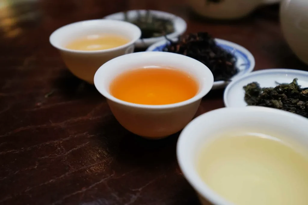 Several ceramic cups hold golden and amber tea on a dark wooden table, surrounded by loose tea leaves, creating a warm, calm tasting scene.
