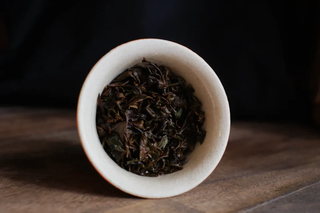 A small white bowl filled with dried tea leaves, placed on a wooden surface with a dark background.