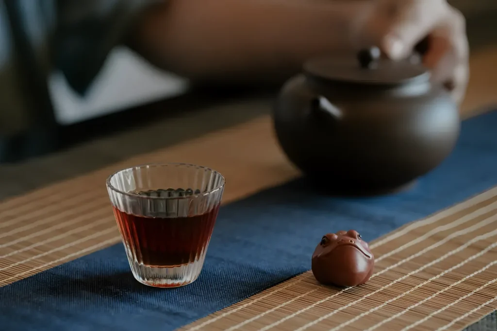 A glass of tea, a teapot, and a small decorative frog figurine on a wooden table with a blue cloth.