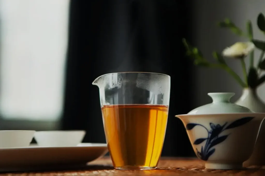 A glass teapot with steaming hot tea, next to a small teacup and a decorative teapot, with a blurred background.