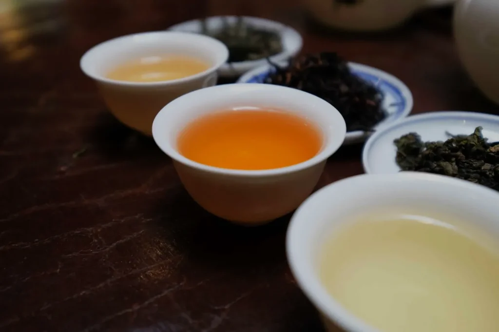 Three small cups of tea in a row, with one cup containing a vibrant orange tea, one with a lighter yellow tea, and a third cup partly obscured. In the background, there are small plates with loose tea leaves.