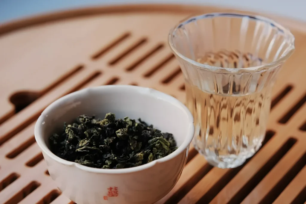 A small bowl of dried green tea leaves next to a clear glass of brewed tea, set on a wooden surface with slatted design.