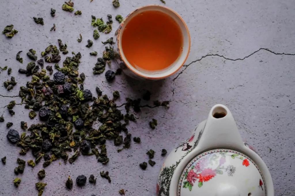 A tea pot, a cup of tea, and dried tea leaves scattered on a textured surface.