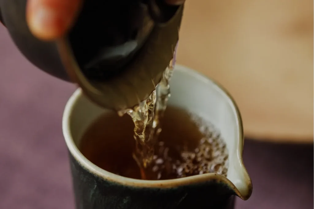 A person pours tea from a teapot into a small cup, with the liquid creating bubbles as it flows.
