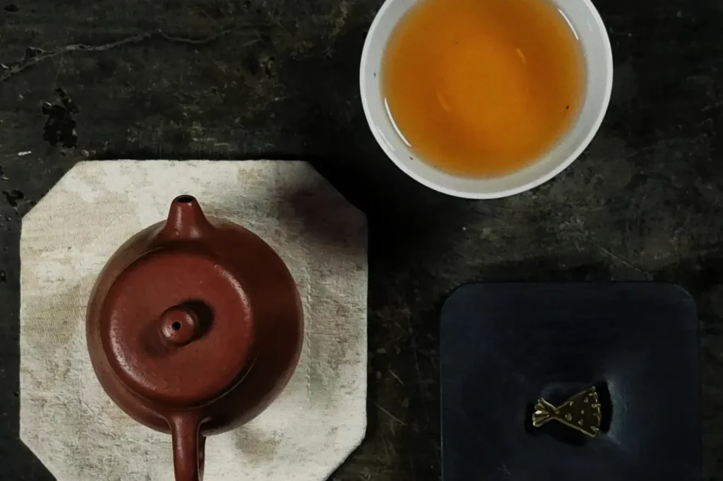A teapot next to a cup of tea on a dark surface.