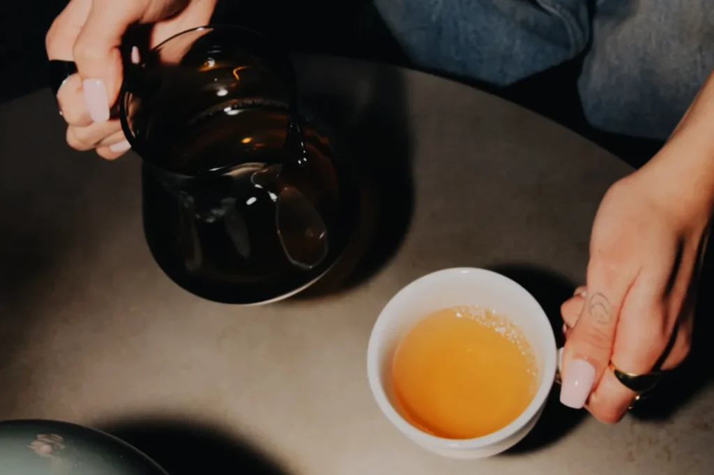 A person is pouring tea from a glass teapot into a small white cup on a table.