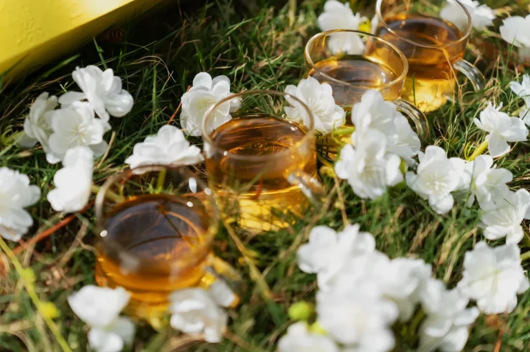 Small glass cups of amber tea nestled among white flowers and green grass in warm sunlight, creating a peaceful, intimate outdoor picnic scene.