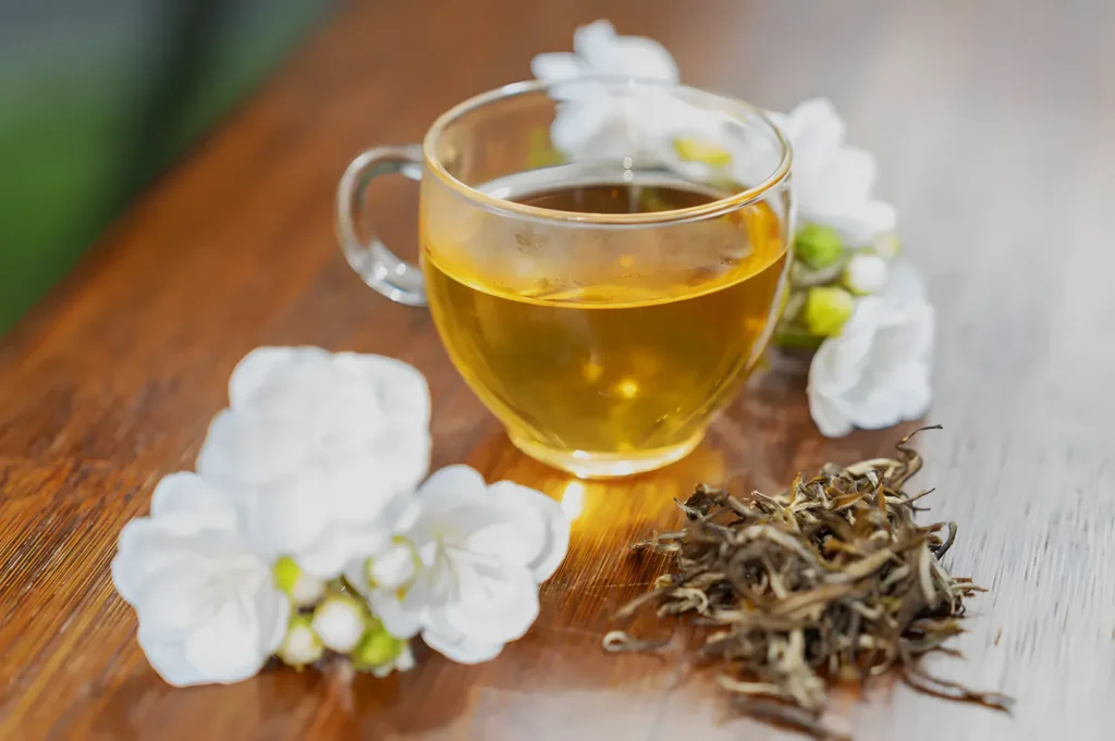 A clear glass cup of golden herbal tea sits on a wooden table beside white blossoms and loose dried herbs, creating a calm, soothing mood.