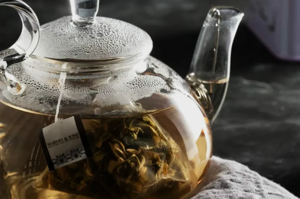 Clear glass teapot filled with amber tea and blooming loose leaves, condensation beading on the lid; warm light creates a calm, cozy mood.
