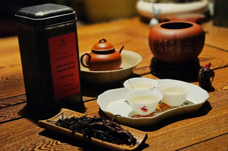 A warmly lit tea setup on a wooden table features a black tea tin, small clay teapot, cups, and a tray of loose leaves, evoking calm tradition.
