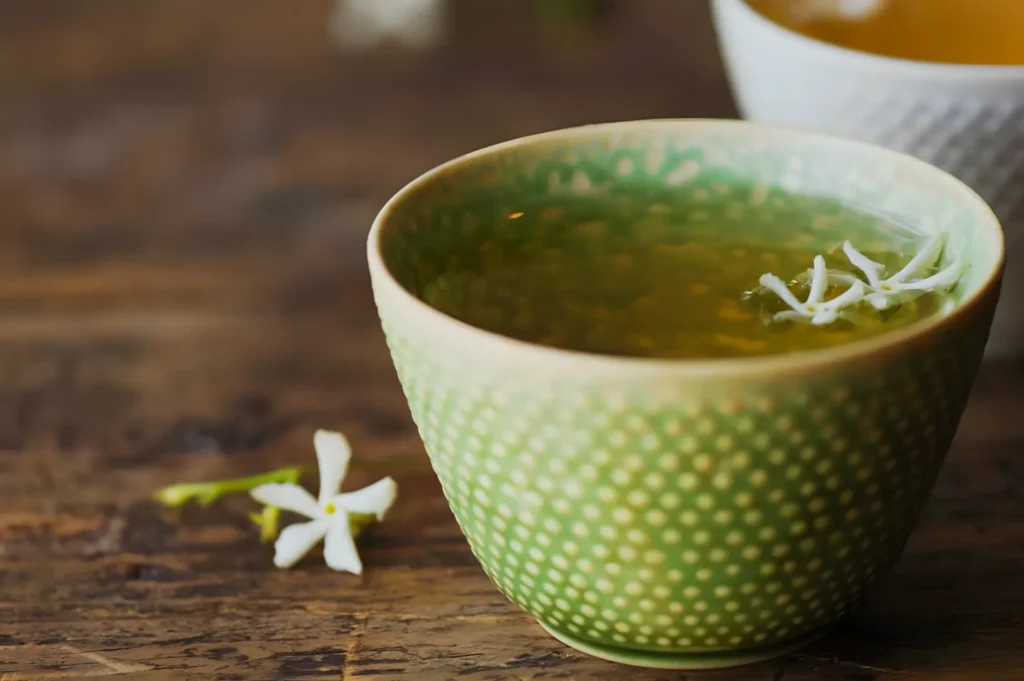 A green patterned ceramic cup of herbal tea, topped with white jasmine blossoms, sits on a rustic wooden table beside a loose flower, creating a calm, cozy mood.