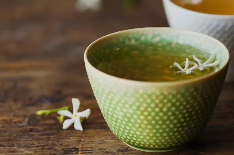 A green patterned ceramic cup of herbal tea, topped with white jasmine blossoms, sits on a rustic wooden table beside a loose flower, creating a calm, cozy mood.