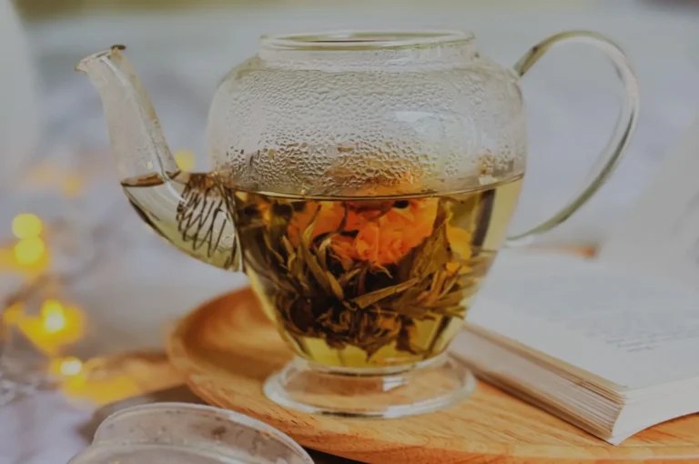 A clear glass teapot steeps orange and yellow blooming tea, with condensation on its lid, resting on a wooden tray beside an open book and warm lights.
