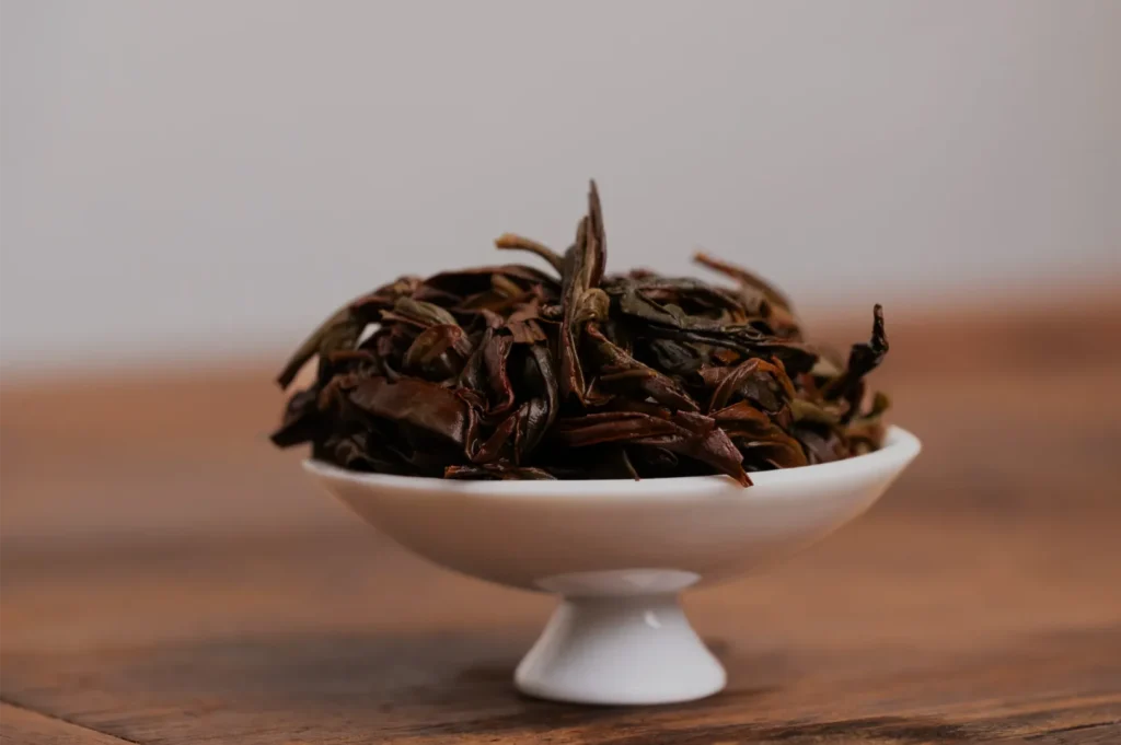 A white pedestal bowl piled with dark, curled loose-leaf tea sits on a wooden table against a plain gray wall, creating a calm, rustic still life.