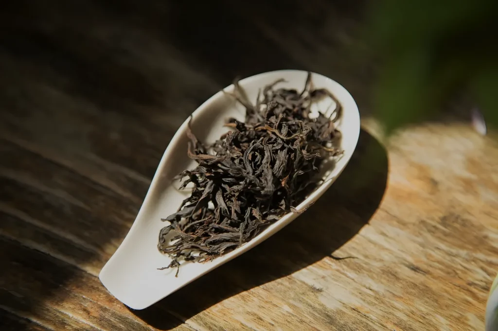 A white ceramic scoop filled with dark, twisted loose tea leaves rests on a sunlit wooden surface, creating a warm, rustic still life.