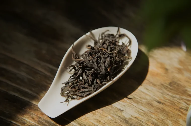 A white ceramic scoop filled with dark, twisted loose tea leaves rests on a sunlit wooden surface, creating a warm, rustic still life.