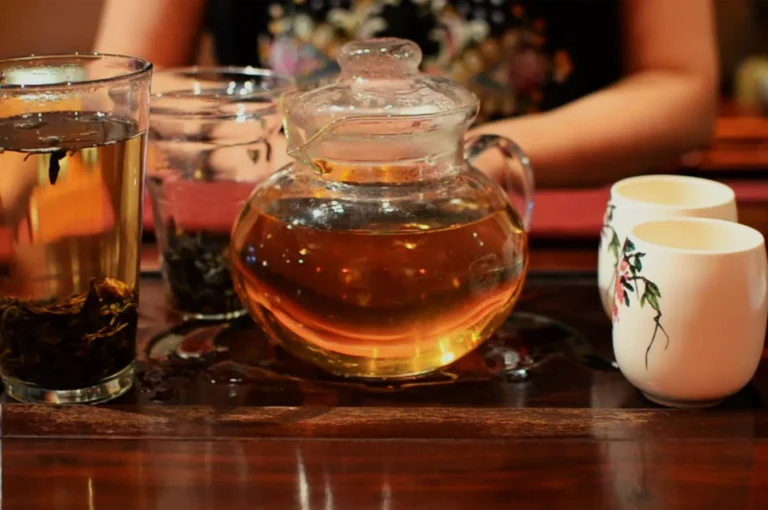 A glass teapot of amber tea sits on a polished wooden table beside tea glasses and floral cups, while a person serves in a warm, relaxed setting.