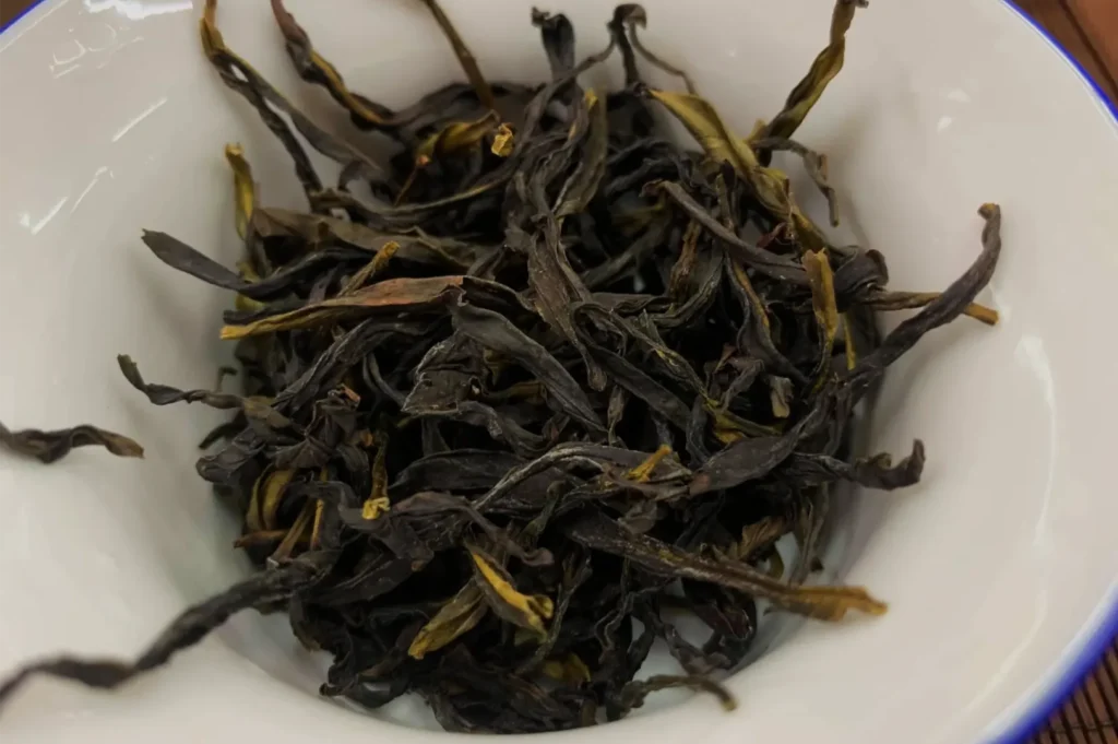 A close-up of dark, twisted loose tea leaves with a few golden tips piled in a white bowl, creating a rustic, earthy impression.