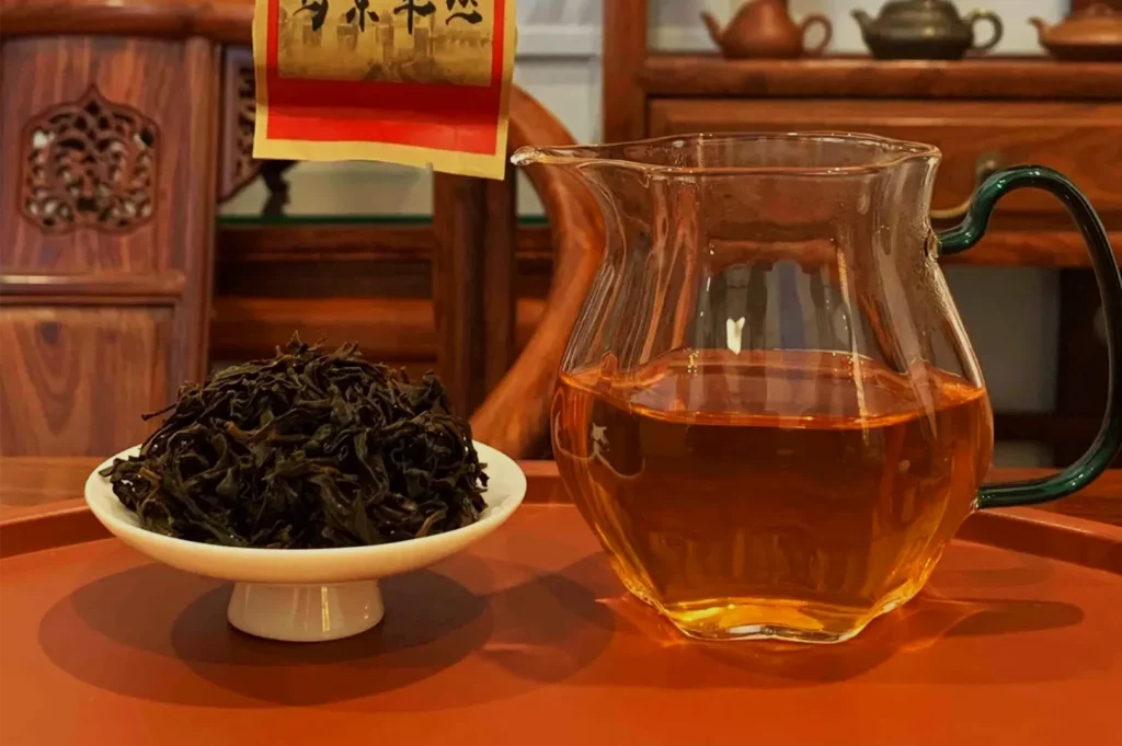 A glass pitcher of amber tea sits beside a dish of loose dark tea leaves on a wooden table, with Chinese tea décor behind them; warm, inviting scene.