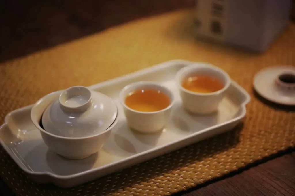 A white ceramic tea set with a lidded pot and two cups of amber tea rests on a tray over a woven placemat, creating a warm, tranquil mood.