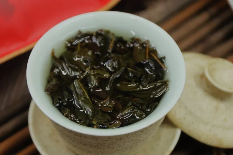 A white bowl filled with dark, glossy loose-leaf tea sits on a wooden tray beside a pale round tea cake, creating a rustic, inviting tea scene.