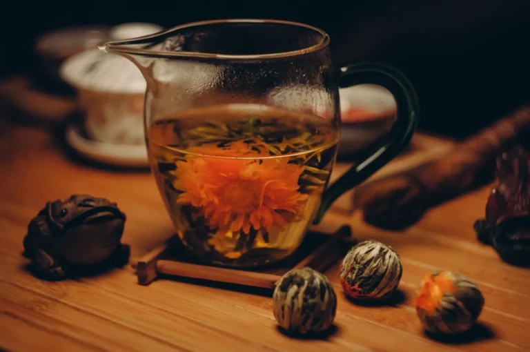 A glass teapot steeps a vivid orange chrysanthemum tea beside dried tea flowers and a dark tea pet on a wooden table, creating a warm, tranquil mood.
