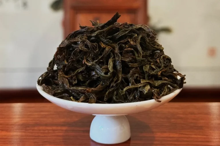 A white pedestal dish piled with dark, twisted dried tea leaves sits on a wooden table, highlighting their rich texture in a calm, rustic setting.