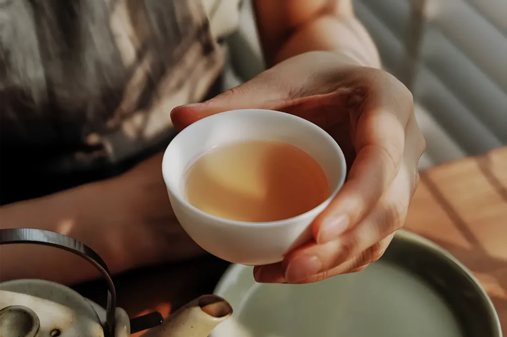 Close-up of a person holding a white cup of amber tea above a kettle, creating a quiet, warm, and comforting moment in a softly lit kitchen.