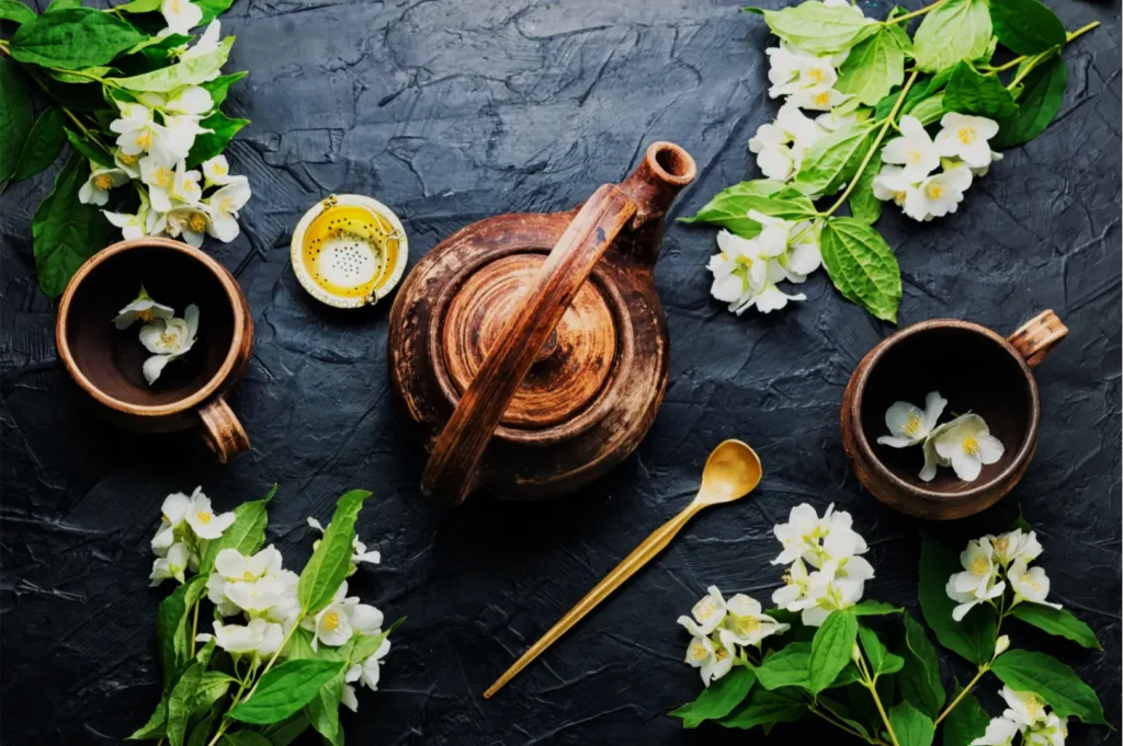 Overhead still life of a wooden mortar and pestle, jasmine flowers, golden herbal tea, and a brass spoon on dark slate, evoking a calm, natural mood.