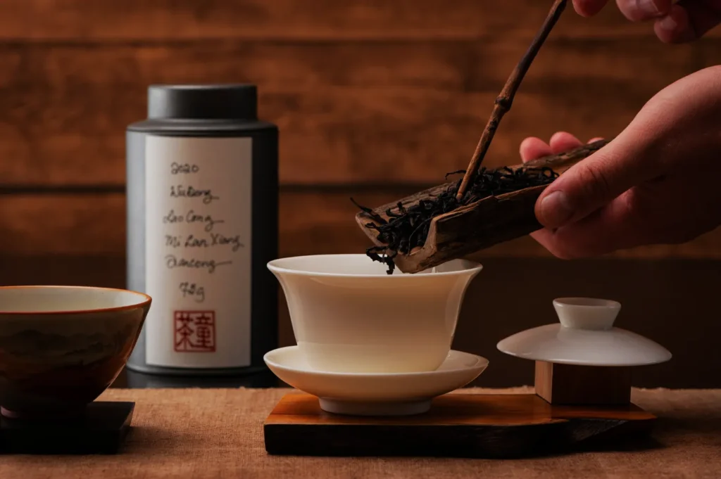 A hand carefully tips dark tea leaves from a wooden scoop into a white porcelain cup, with a tea tin and bowls on a warm wooden table.