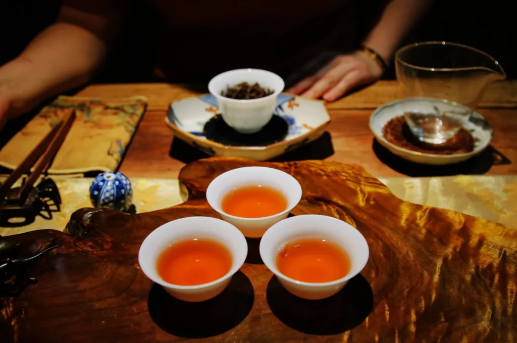 Three white cups of amber tea sit on a wooden tray, with a teapot and tea leaves behind them; hands frame a quiet tea ceremony.