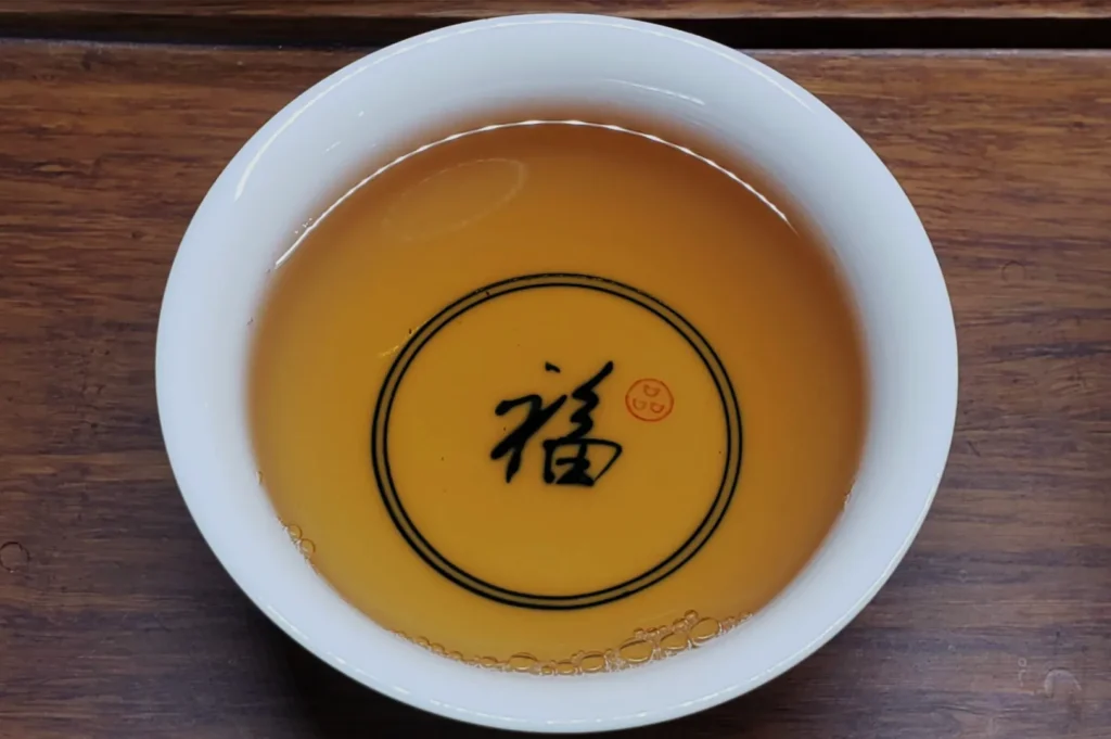 Overhead view of amber tea in a white bowl on a wooden table, decorated with black Chinese calligraphy and a red seal; calm, traditional mood.